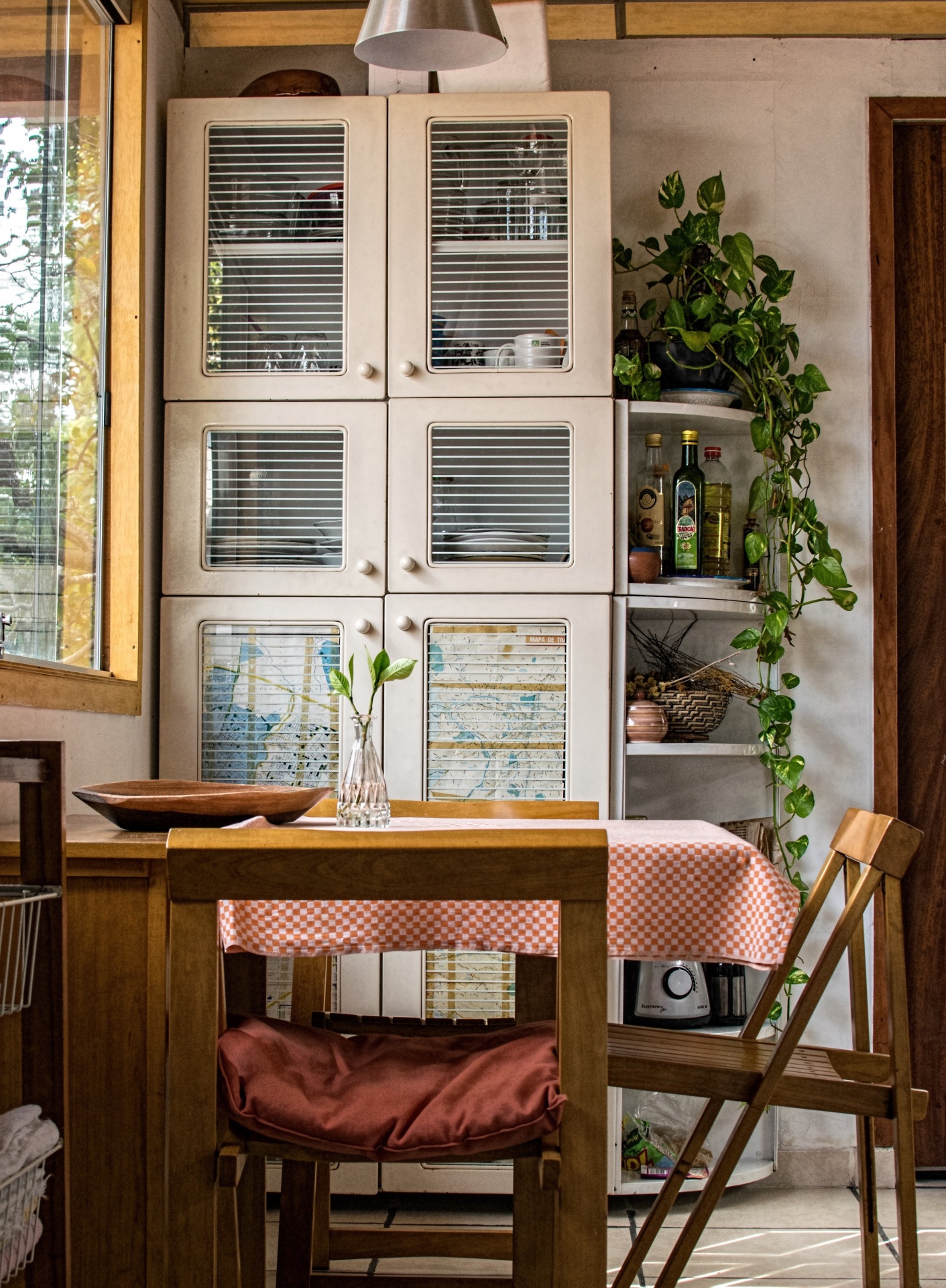 Móvel de cozinha em madeira clara com portas de vidro, mesa com toalha xadrez e cadeira com almofada