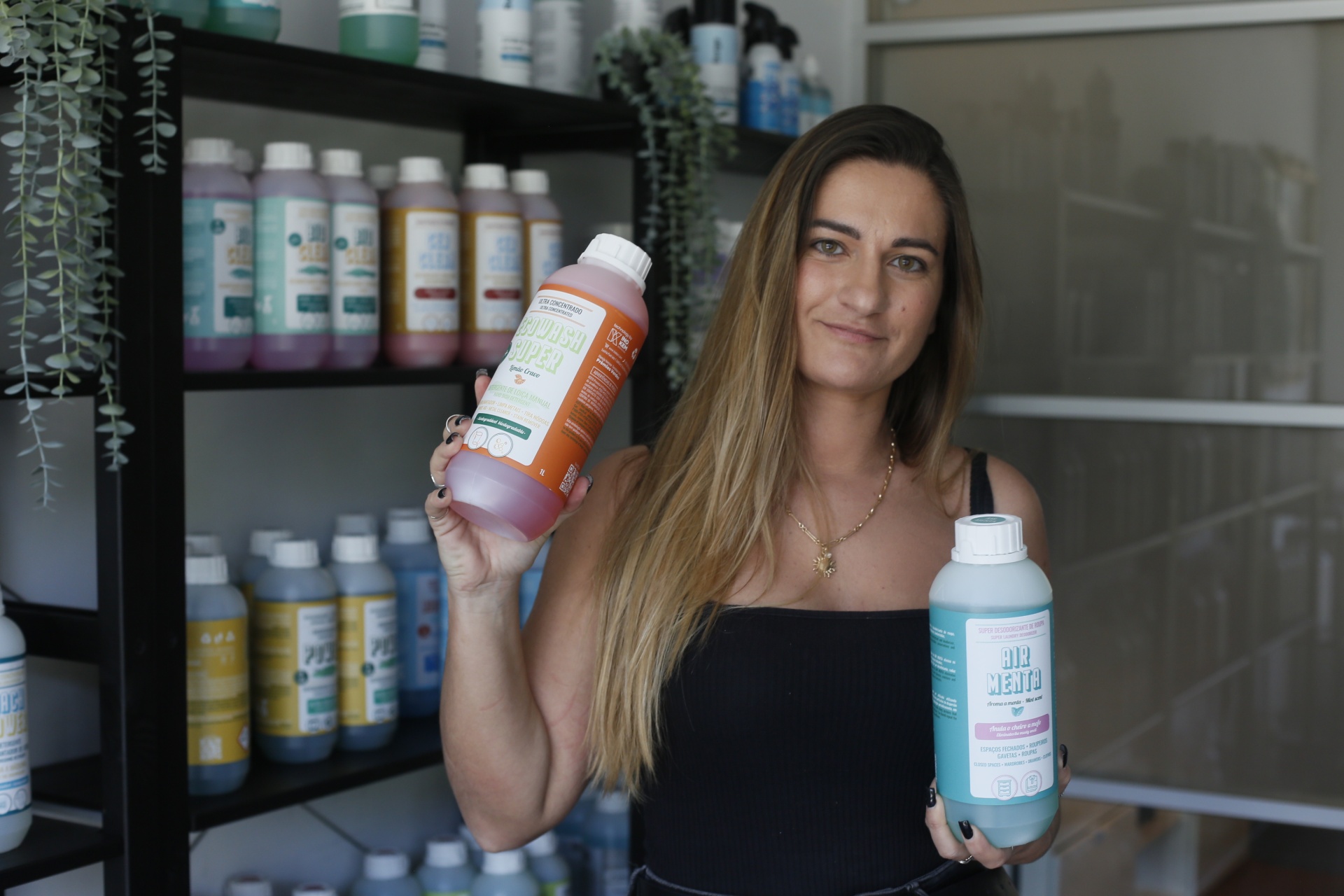 Mulher segura duas garrafas de produtos coloridos em frente a estantes com mais garrafas.