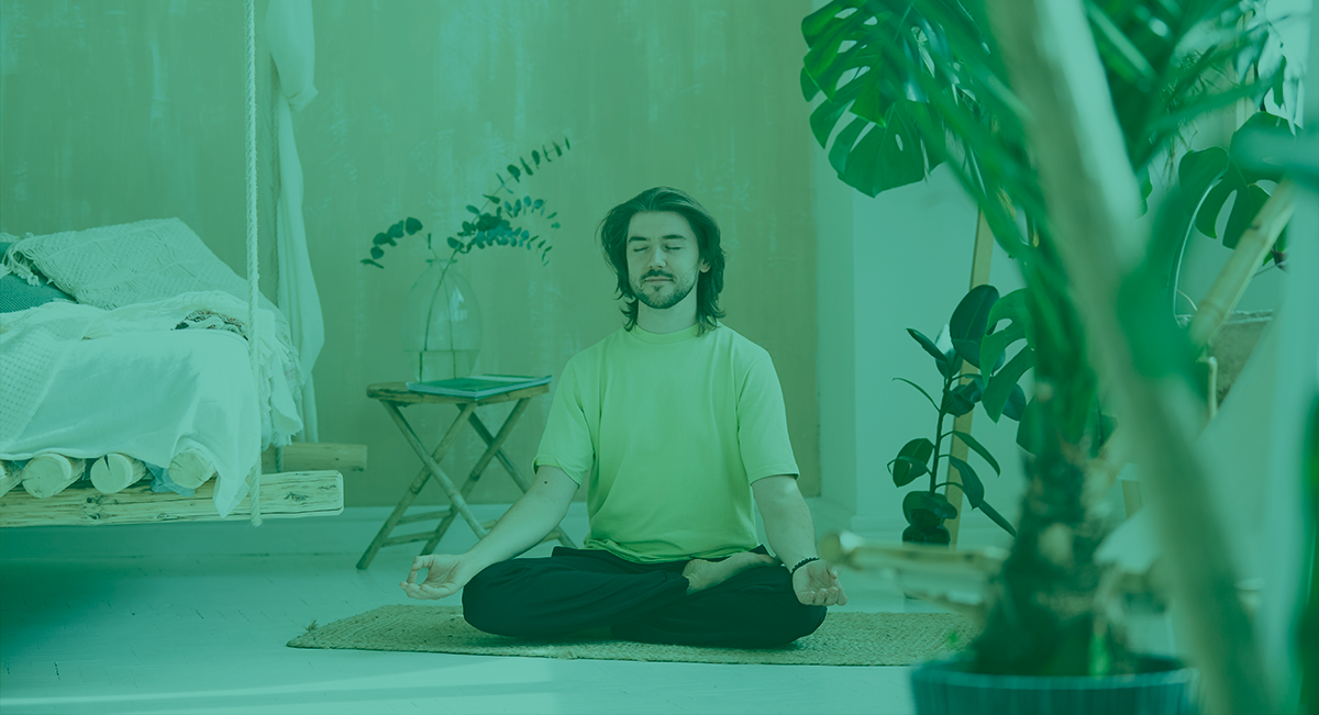 Como é que a limpeza de casa ajuda na saúde e bem-estar? Homem sentado de pernas cruzadas em quarto verde com plantas e cama branca