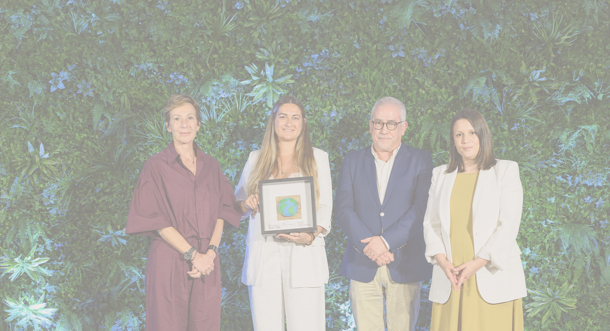 Grupo de quatro pessoas em roupa formal com fundo verde de plantas, uma pessoa segura um quadro com desenho do globo terrestre.