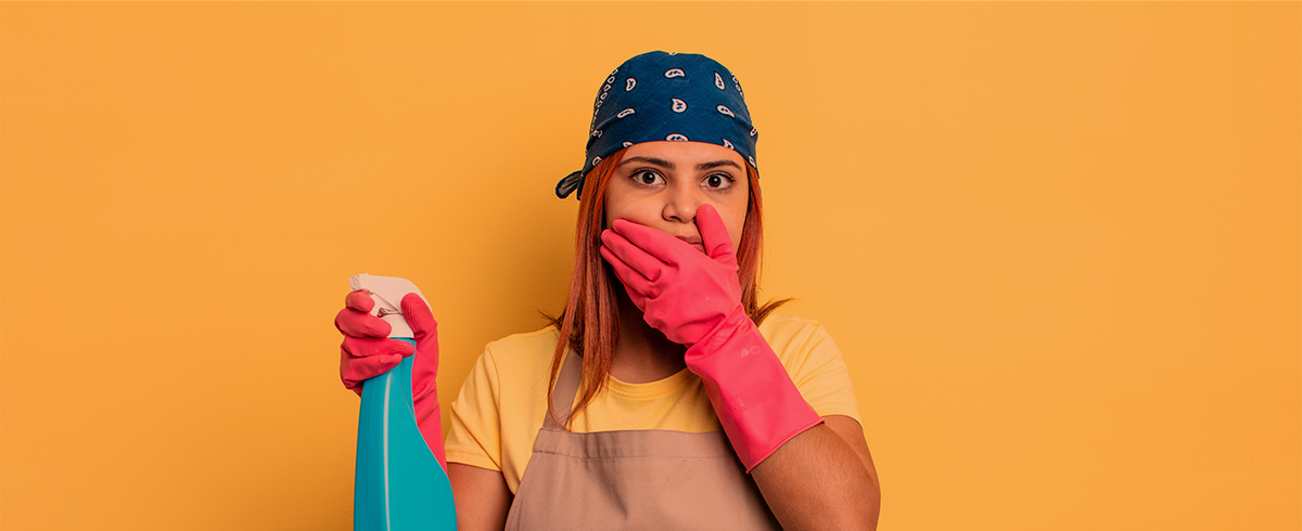 Mulher com luvas rosa e bandana azul segurando frasco spray azul com fundo laranja