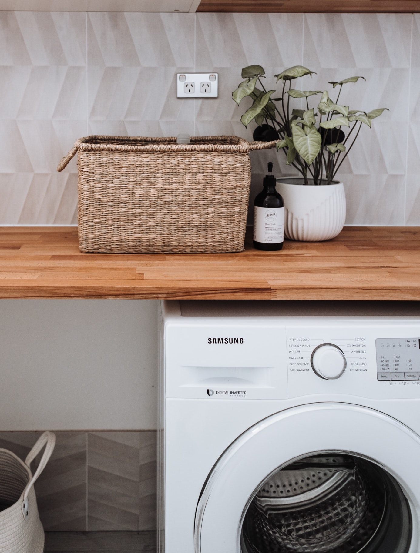 Máquina de lavar branca Samsung com cesta de vime e planta numa bancada de madeira