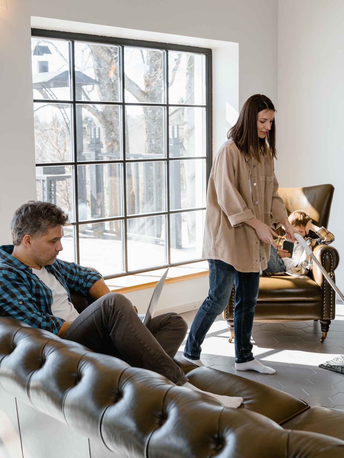 Sala de estar com sofá castanho, poltrona, homem com portátil, mulher a aspirar e criança na poltrona