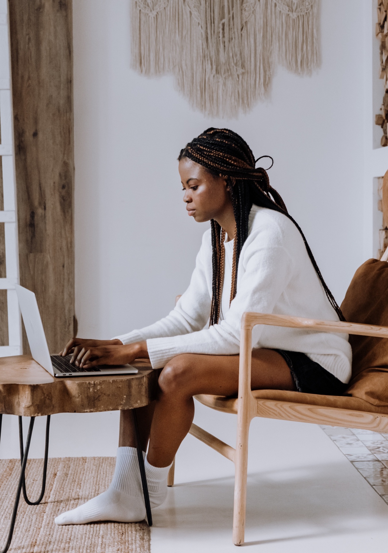 Mulher com camisola branca e meias a usar portátil numa cadeira de madeira com mesa de madeira