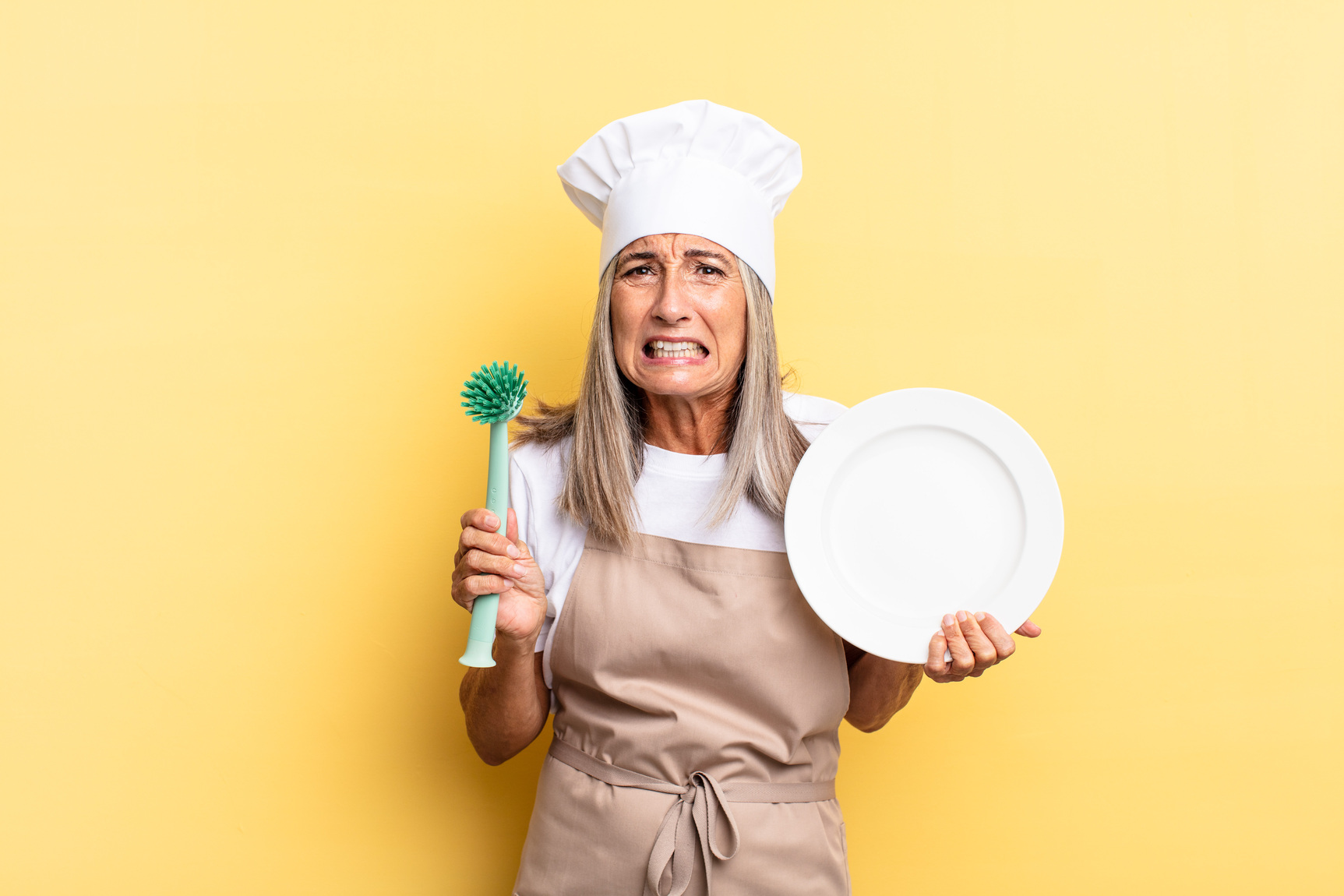 Mulher com avental bege e chapéu de chef segurando escova de pratos e prato branco em fundo amarelo