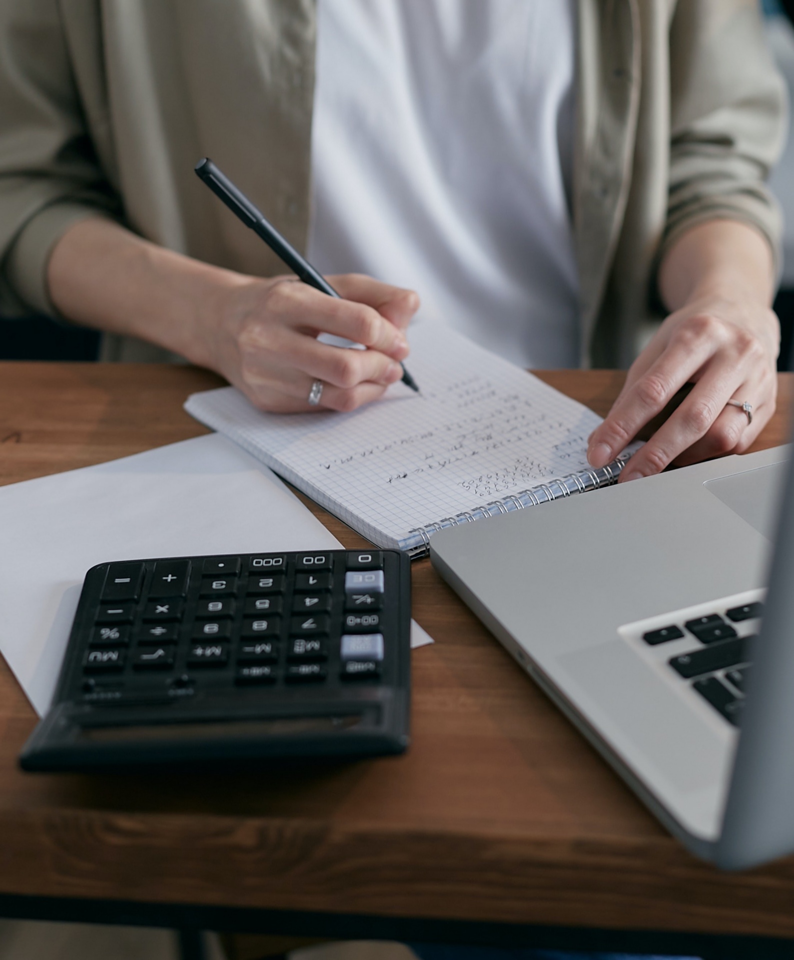 Como poupar em tempos de crise? Pessoa a escrever num caderno ao lado de computador portátil e calculadora numa mesa de madeira