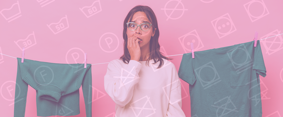 Símbolos de lavagem da roupa: o que querem dizer? Mulher com óculos entre duas roupas verdes penduradas numa corda de estender roupa com fundo cor de rosa