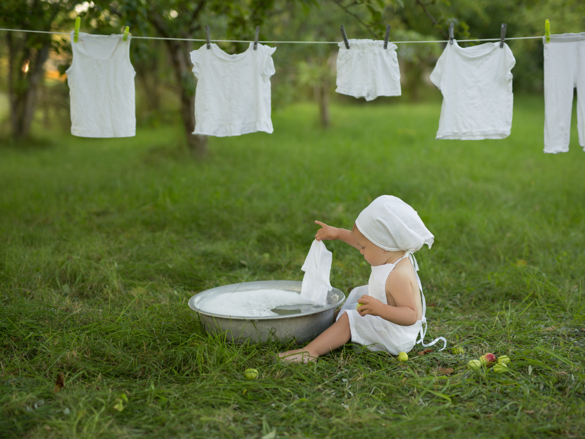 Criança vestida de branco com lenço na cabeça a brincar com roupa branca e bacia metálica ao ar livre