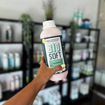 Frasco plástico rosa com rótulo BIO SOFT segurado em frente a prateleiras com produtos.