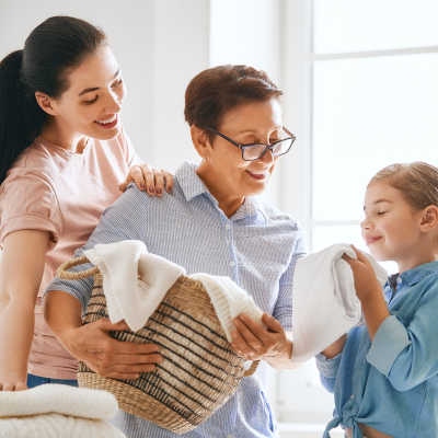 Três mulheres a manusear roupa e uma cesta de roupa em ambiente doméstico claro com plantas