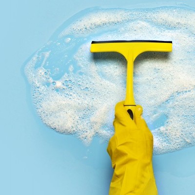 Mão com luva amarela segurando limpa-vidros amarelo numa superfície azul com espuma branca