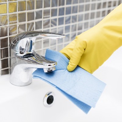 Mão com luva amarela a limpar torneira cromada com pano azul numa pia branca