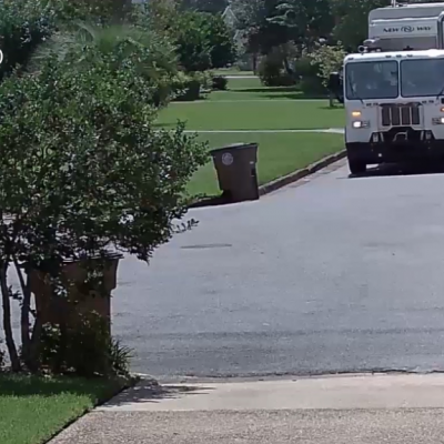 Camião de recolha de lixo branco numa rua residencial com relva e lixeiras castanhas