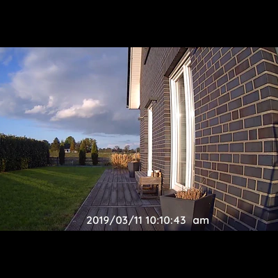 Casa em tijolo escuro com varanda de madeira e cadeira com almofada, relvado e céu azul com nuvens.
