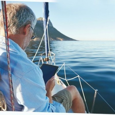 Homem sentado num veleiro a ler um livro com mar e montanha ao fundo