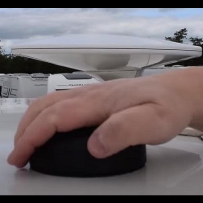 Mão sobre dispositivo preto arredondado em superfície branca com veículos e estrutura branca ao fundo