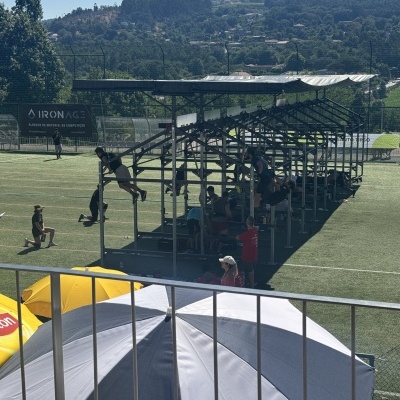 Pessoas a treinar numa estrutura de treino num campo desportivo com guarda-sóis e montanhas ao fundo