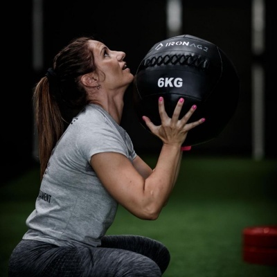 Mulher segurando bola medicinal preta com texto 'IRON AG3' e '6KG' num ginásio