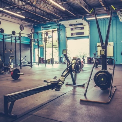Ginásio com equipamentos de remo, barras e pesos, parede azul e luz natural