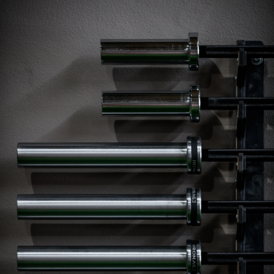 Cinco barras olímpicas de musculação cromadas organizadas num suporte preto junto a uma parede cinzenta