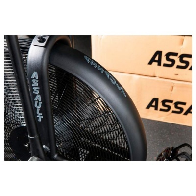 Bicicleta estática preta com texto 'ASSAULT' azul clara no aro e caixas de embalagem ao fundo.