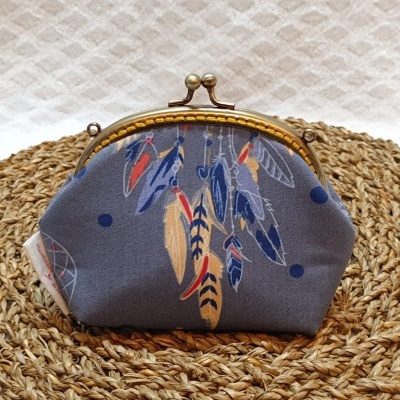 Carteira pequena azul com padrão de penas coloridas e fecho metálico dourado em superfície de palha