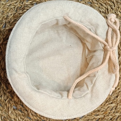 Chapéu de tecido castanho claro com cordão e pompom sobre superfície de ráfia entrelaçada