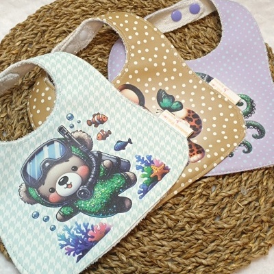 Três babetes para bebé com estampados coloridos e diferentes padrões de bolinhas, sobre tapete trançado castanho.