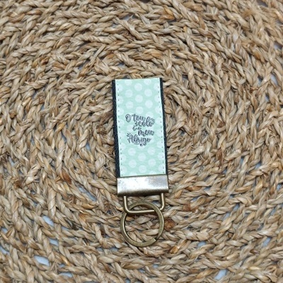 Chaveiro verde claro com bolinhas brancas e texto com argola dourada sobre tapete de fibras naturais
