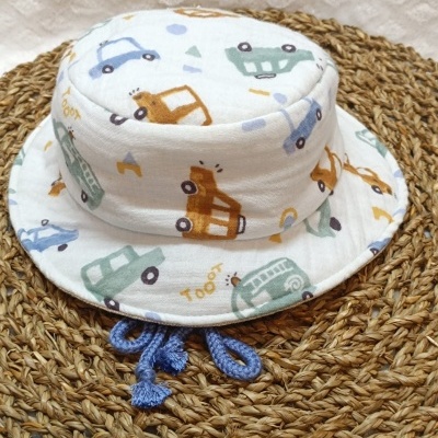 Chapéu para bebé branco com carrinhos coloridos e cordões azuis sobre tapete de fibras.