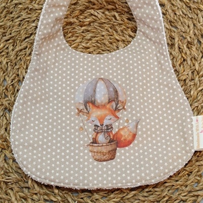 Babete bege com padrão de estrelas brancas e raposa num balão, com fecho botão branco