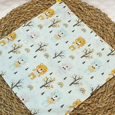 Pano de cozinha azul claro com desenhos de ursos e árvores, com alça amarela, sobre base de fibra natural
