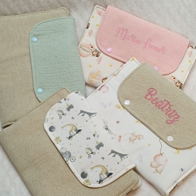 Capas de tecido para livros com padrões infantis e nomes bordados colocadas sobre uma superfície macia clara.