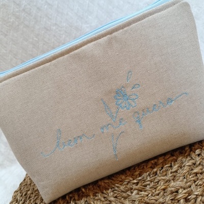 Necessaire bege com bordado azul bem me quero e flor, em fundo branco e palha trançada.