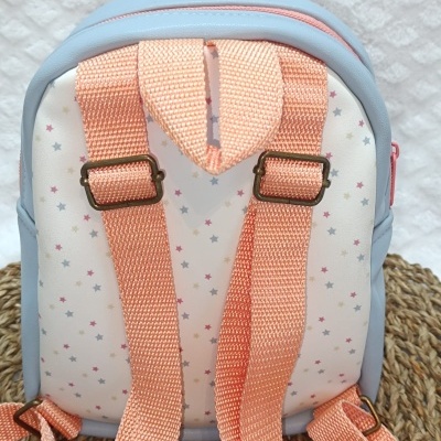 Mochila pequena branca com estrelas coloridas e alças laranja sobre superfície de fibras naturais.