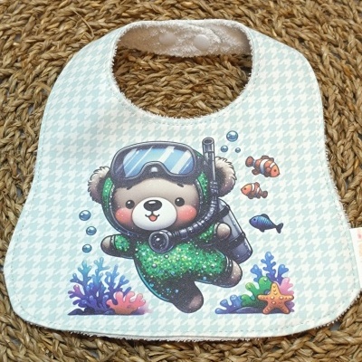 Babete de bebé azul claro com padrão pied-de-poule e estampa de urso mergulhador com peixes e coral.