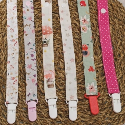 Seis fitas para chupeta em tecido com padrões diversos e fechos plásticos em diferentes cores, dispostas sobre base trançada.
