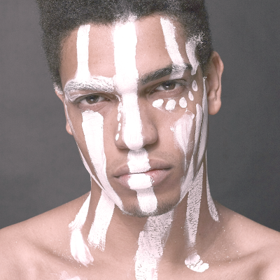 Jovem com pinturas brancas tribais no rosto e pescoço em fundo cinzento.