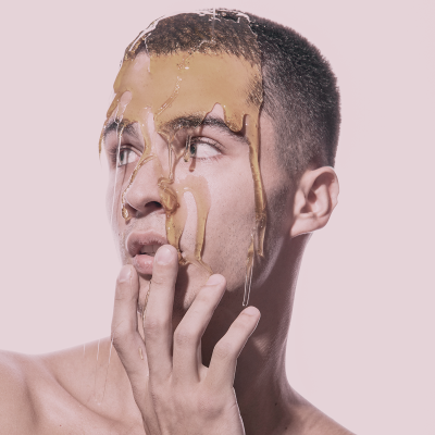 Homem jovem com líquido cor de caramelo no rosto e fundo rosa claro.