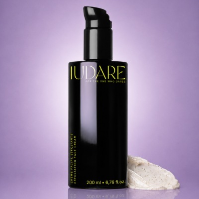 Frasco preto de creme facial esfoliante com textura cremosa branca ao lado, fundo roxo.