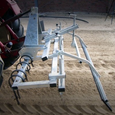 Equipamento agrícola metálico preso a trator sobre areia em ambiente fechado.