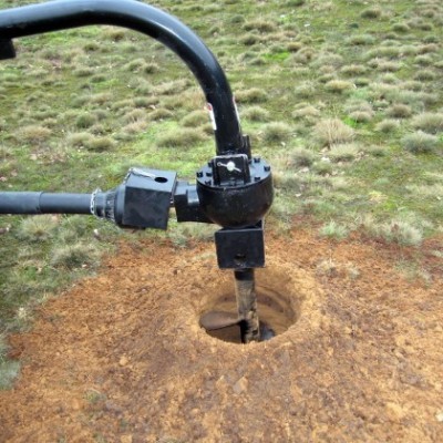 Equipamento de perfuração metálico preto perfurando solo avermelhado em área com erva