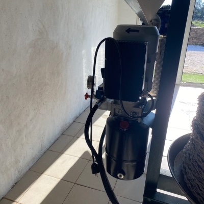 Máquina industrial preto e cinzenta com funil e cabos junto a parede branca e chão de azulejos