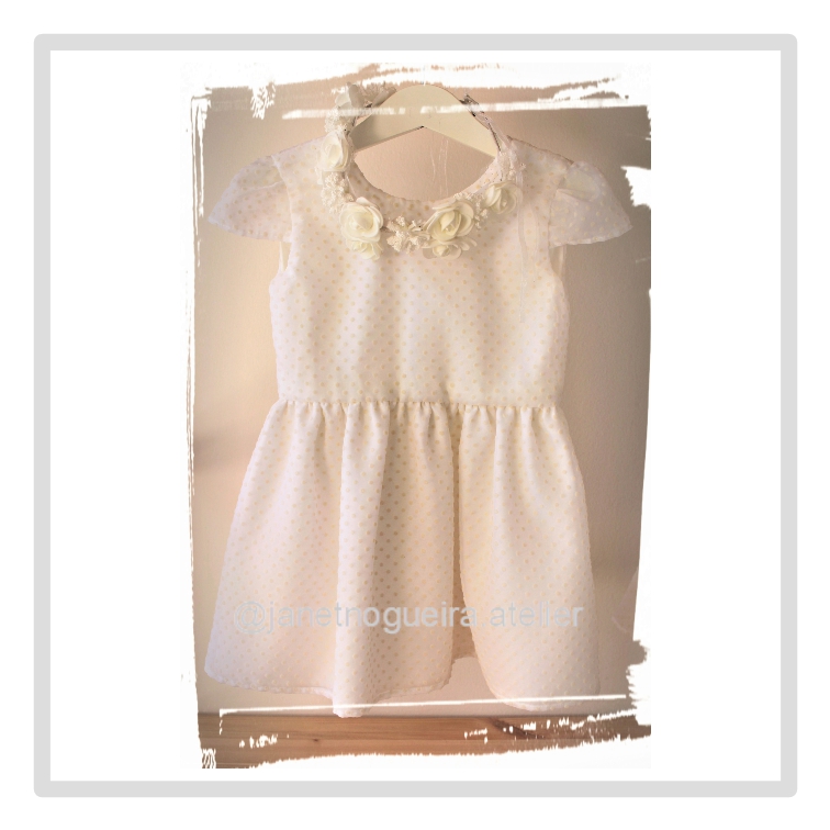 Vestido Violeta Vestido branco infantil com flores e pérolas no decote, pendurado em cabide.