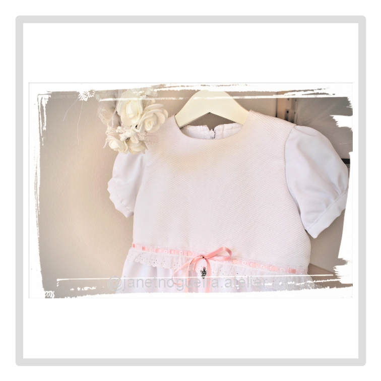 Vestido Matilde Vestido branco de bebé com fita rosa e mangas franzidas pendurado num cabide