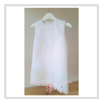 Vestido branco de criança em textura fina, com renda e laço rosa na barra, pendurado num cabide.