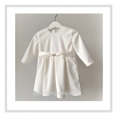 Vestido branco de manga comprida para criança com faixa e laço prateado pendurado num cabide branco.