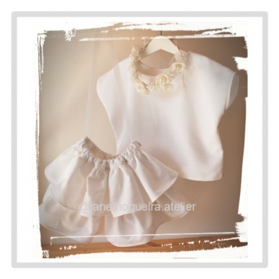 Conjunto de roupa infantil branco com blusa e saia folhos pendurados em suporte de madeira