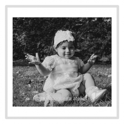 Bebé em vestido branco rendilhado com faixa e sapatinhos sentada no relvado ao ar livre