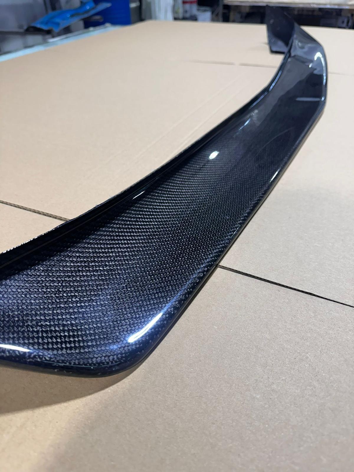 Spoiler de automóvel em fibra de carbono preta com textura visível, sobre cartão castanho